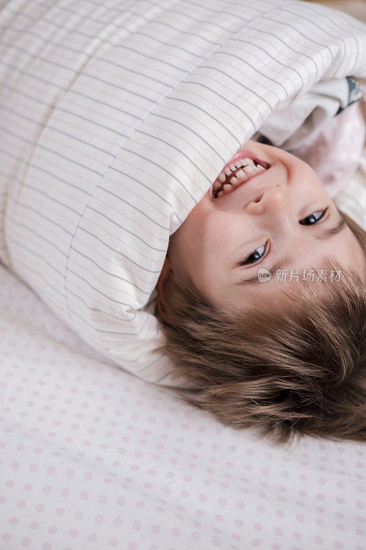Child toddler boy is wrapped in a blanket like a roll, his head sticks out of the blanket. He is laughing, having fun on the bed.